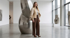 A woman stands in a modern art gallery wearing a beige cropped jacket, a light top, brown wide-leg trousers, and brown heels, positioned in front of a large metallic sculpture.