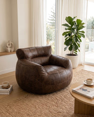A round, low-profile brown leather armchair with a distressed finish, styled in a bright, modern living room next to a potted fiddle-leaf fig plant and a small wooden coffee table.