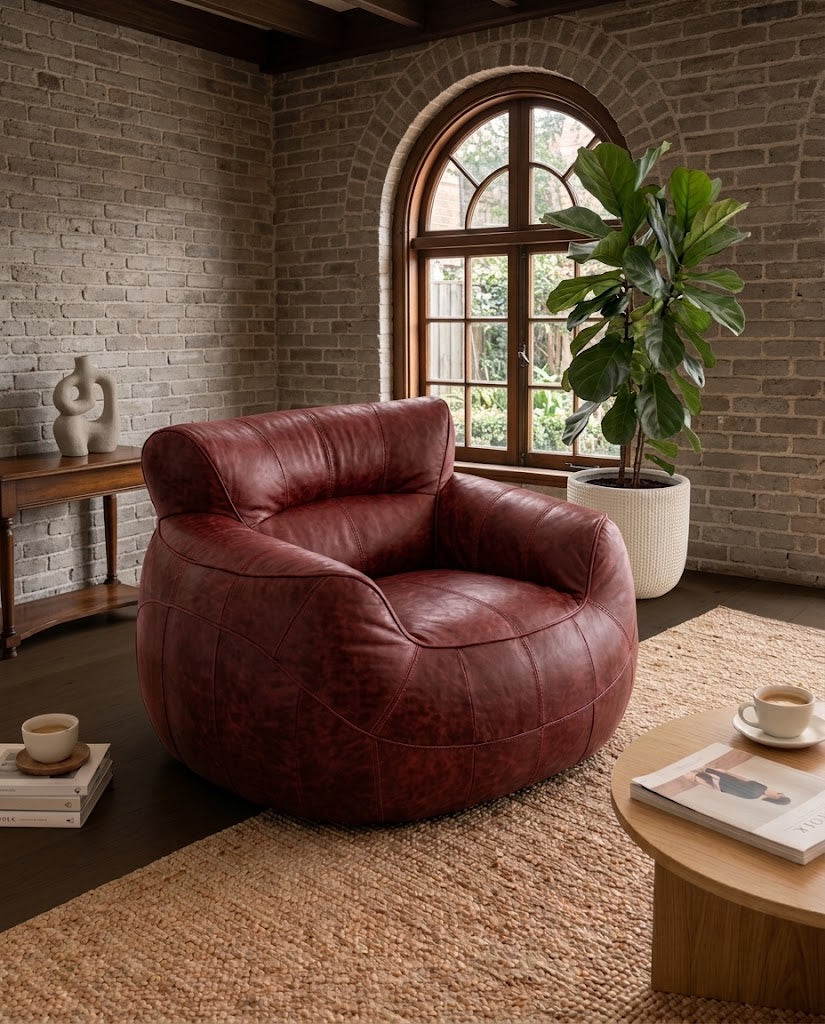 A round, low-profile leather armchair in a distressed reddish-brown color, styled in a rustic living room with a brick wall, arched window, potted plant, and wooden coffee table.