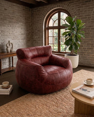 A round, low-profile leather armchair in a distressed reddish-brown color, styled in a rustic living room with a brick wall, arched window, potted plant, and wooden coffee table.