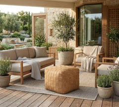 Outdoor patio furniture set with a light wood frame, beige cushions, a woven tan pouf, and neutral throw blankets, arranged on a rug over a wooden deck with potted plants and a stone wall background.