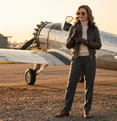 A woman stands on an airfield runway in front of a vintage silver airplane, wearing a dark brown leather jacket, dark gray trousers, brown leather gloves, and sunglasses.