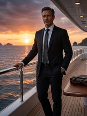 A man in a dark charcoal suit, white dress shirt, and black tie stands on a yacht deck at sunset, leaning on a railing with the ocean and sky behind him.