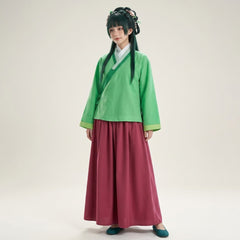 A model wearing a traditional Chinese Hanfu outfit consisting of a green short-sleeved top with a diagonal collar, a deep magenta wide-leg skirt, and teal shoes, standing against a plain light beige studio background.