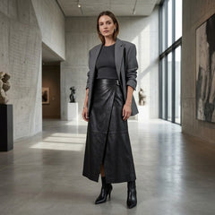 A woman stands in a modern concrete gallery wearing a dark gray blazer, black top, black leather wrap skirt, and black ankle boots.