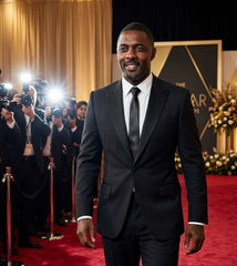 A man in a black suit, white dress shirt, and black tie walking on a red carpet at an awards show, with photographers and stage lighting visible in the background.