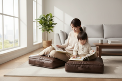 A woman and a young child sit on brown floor cushions in a bright, modern living room, reading books together. The scene includes a light gray sofa, a wooden side table, a potted plant, and large windows with soft natural light.