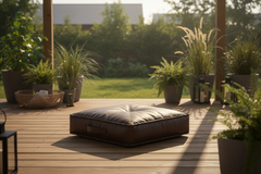 A square brown leather floor cushion sits on a wooden deck in a backyard patio setting with potted plants and a blurred green lawn in the background.