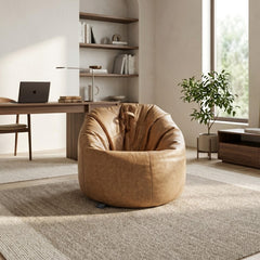 A round tan leather bean bag chair in a modern, sunlit home office setting with a desk, laptop, bookshelves, and a potted plant.