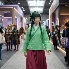 A woman wearing a detailed cosplay outfit stands in a convention center aisle. The outfit features a light green top with a darker green V-neck trim, a white collar, and a dark red wide-leg skirt.