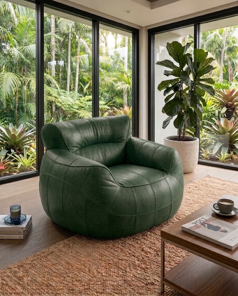 A modern round green leather armchair in a bright, contemporary room with large windows overlooking a lush tropical garden. A wooden coffee table, a woven rug, and a potted fiddle-leaf fig plant are visible in the foreground.