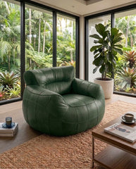 A modern round green leather armchair in a bright, contemporary room with large windows overlooking a lush tropical garden. A wooden coffee table, a woven rug, and a potted fiddle-leaf fig plant are visible in the foreground.