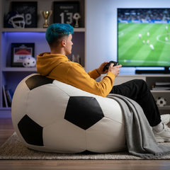A man sits on a giant soccer ball bean bag chair in a living room, holding a game controller in front of a TV showing a soccer match.