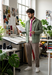 A man in a creative studio stands at an drafting table holding a sheet of paper, wearing a sage green overshirt, pink tie, beige trousers, and white sneakers. The room is filled with plants, color swatches, and design supplies.