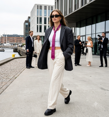 A woman walking outdoors in a city near the water, wearing a dark blazer, white cropped top, white wide-leg trousers, a bright magenta tie, and black sunglasses.