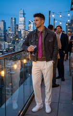 A man standing on a rooftop terrace at dusk wearing a dark brown jacket, a bright pink tie, cream trousers, and white sneakers, holding a cocktail glass. City skyline lights are visible in the background.