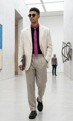 A man walking through a modern art gallery wearing a cream-colored linen blazer, beige trousers, a black shirt, and a bright magenta tie.