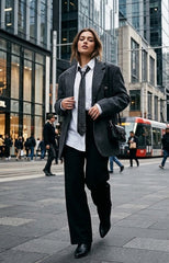 A woman walking on a city street wearing a dark gray oversized blazer, white shirt, black tie, black wide-leg trousers, and black boots, with a black shoulder bag.